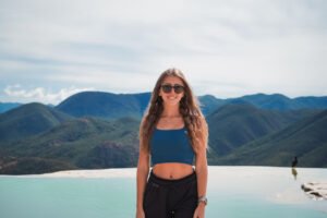 A woman in a dark crop top sitting on a stone wall with a scenic mountain range in the background