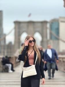 A woman with sunglasses walking outdoors with the Brooklyn Bridge visible in the background