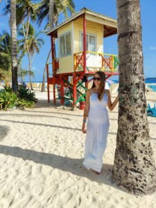 Woman in a white maxi dress standing by a palm tree on a tropical beach near a lifeguard tower