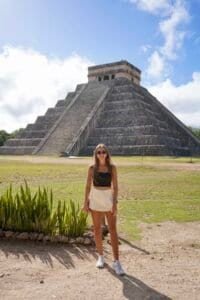 A wide shot of a woman standing before the massive ancient pyramid of Chichen Itza